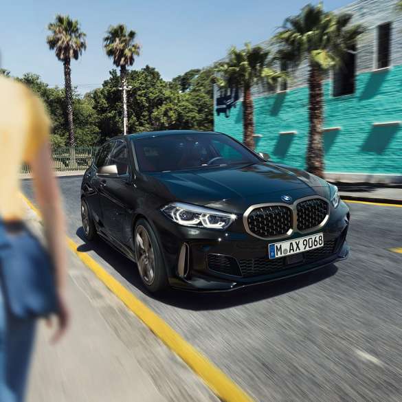 BMW M135i xDrive, Dreiviertel-Frontansicht. BMW M135i xDrive in Saphirschwarz metallic, Dreiviertel-Frontansicht fahrend mit Frau im Vordergrund.