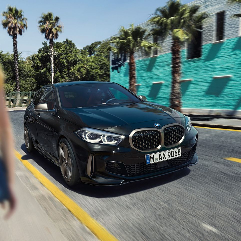 BMW M135i xDrive, Dreiviertel-Frontansicht. BMW M135i xDrive in Saphirschwarz metallic, Dreiviertel-Frontansicht fahrend mit Frau im Vordergrund.