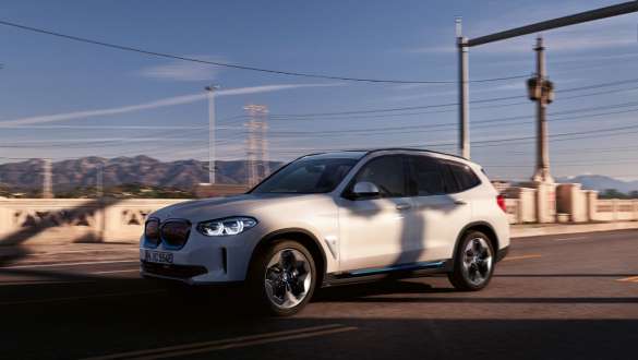 Adaptives M Fahrwerk BMW iX3 G08 fahrend auf Straße Adaptives M Fahrwerk BMW iX3 G08 2020 Mineralweiß metallic Dreiviertel-Frontansicht fahrend auf Straße