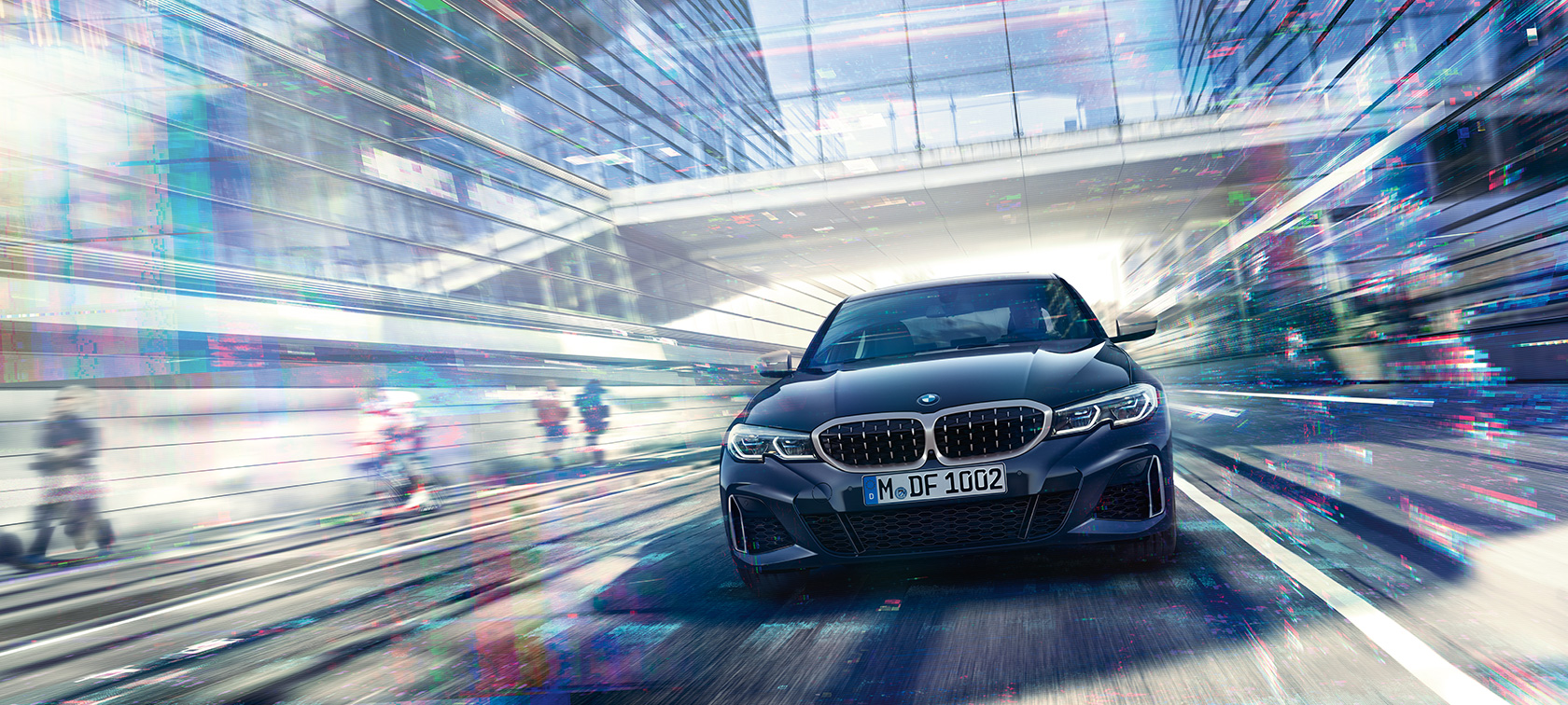 BMW 3er Limousine, Frontalaufnahme BMW M340i xDrive. Frontalaufnahme der fahrenden BMW M340i xDrive Limousine zwischen urbanen Glasgebäuden.