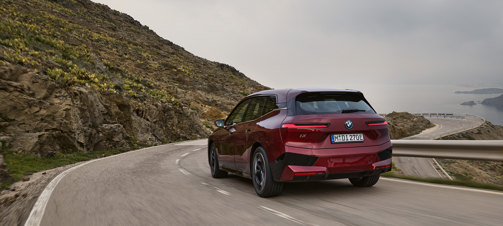 BMW iX i20 2021 elektrischer SUV BMW iX xDrive50 fahrend auf Küstenstraße BMW iX i20 2021 elektrischer SUV BMW iX xDrive50 Aventurinrot Heckansicht fahrend auf Küstenstraße