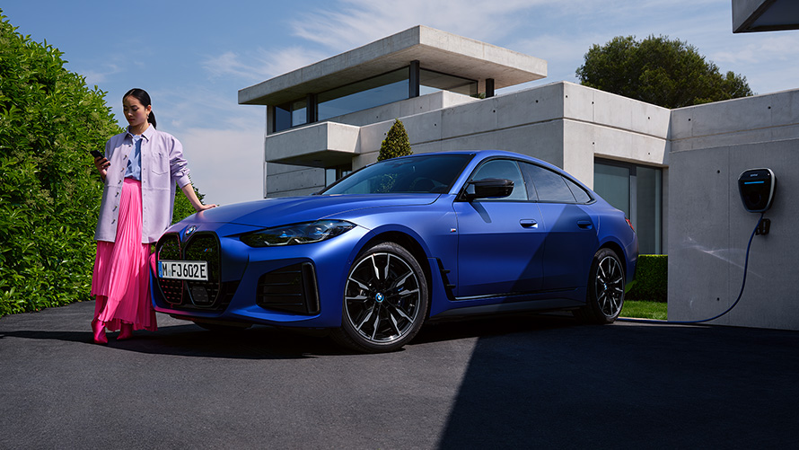 BMW i4 M50 G26 2021 BMW Individual Frozen Portimao Blau metallic Dreiviertel-Frontansicht stehend mit Frau vor Haus