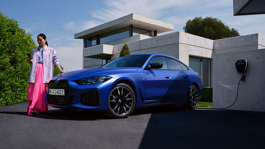 Dreiviertel-Seitenansicht BMW i4 M50 G26 2021 mit Frau vor Haus BMW i4 M50 G26 2021 BMW Individual Frozen Portimao Blau metallic Dreiviertel-Seitenansicht mit Frau vor Haus