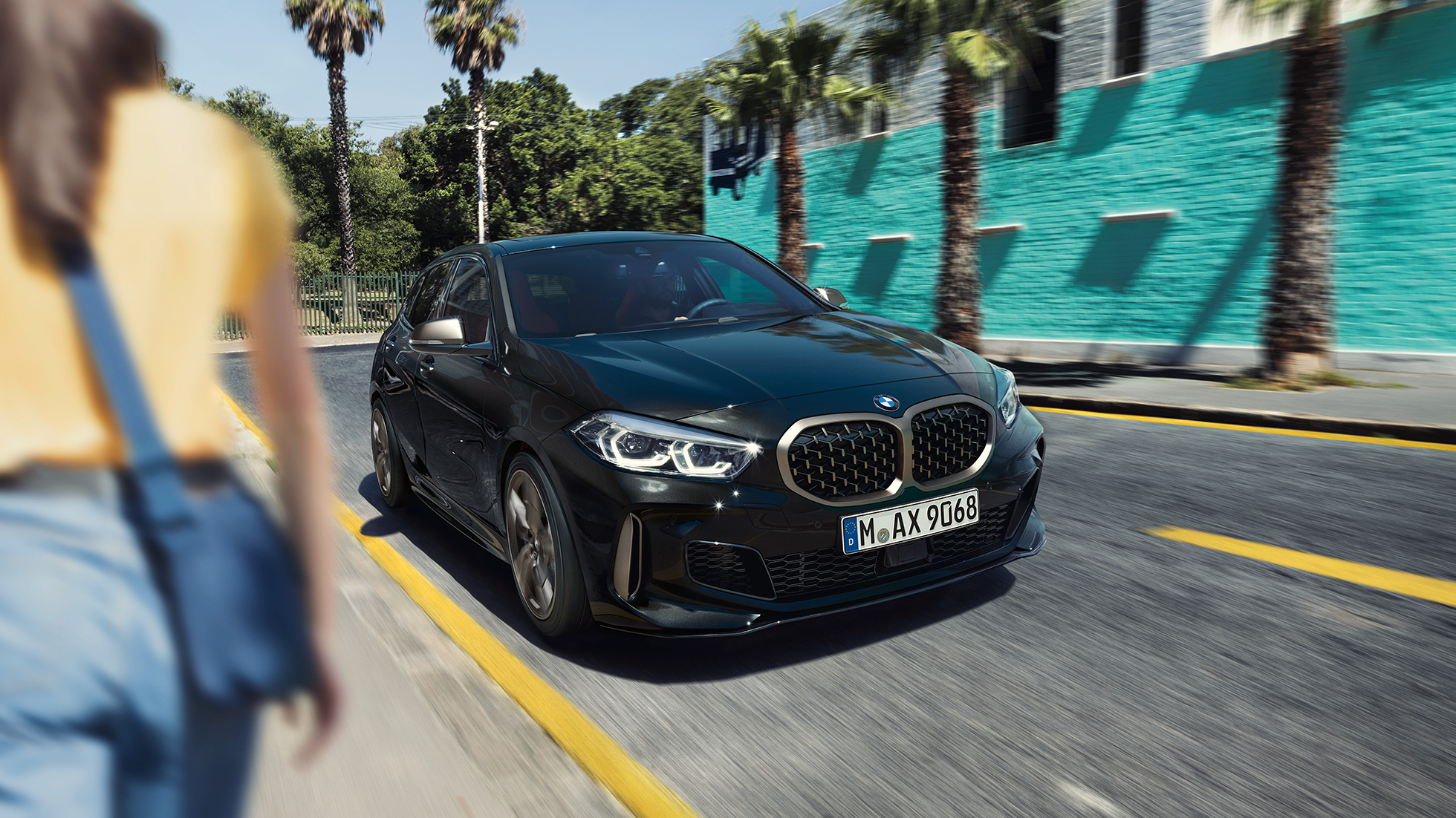 BMW M135i xDrive, Dreiviertel-Frontansicht. BMW M135i xDrive in Saphirschwarz metallic, Dreiviertel-Frontansicht fahrend mit Frau im Vordergrund.