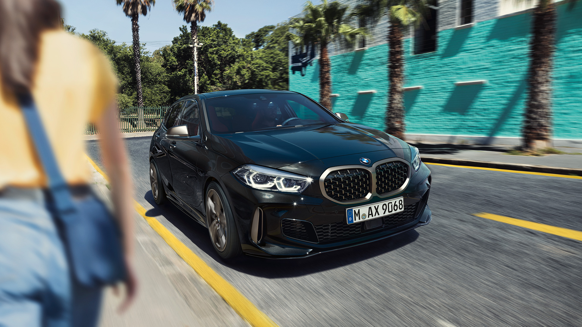 BMW M135i xDrive, Dreiviertel-Frontansicht. BMW M135i xDrive in Saphirschwarz metallic, Dreiviertel-Frontansicht fahrend mit Frau im Vordergrund.