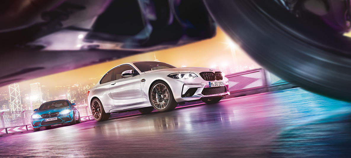 BMW M2 Competition auf spiegelndem Asphalt Zwei BMW M2 Competition auf Rennstrecke vor nächtlicher Großstadtkulisse aus Bodenperspektive.