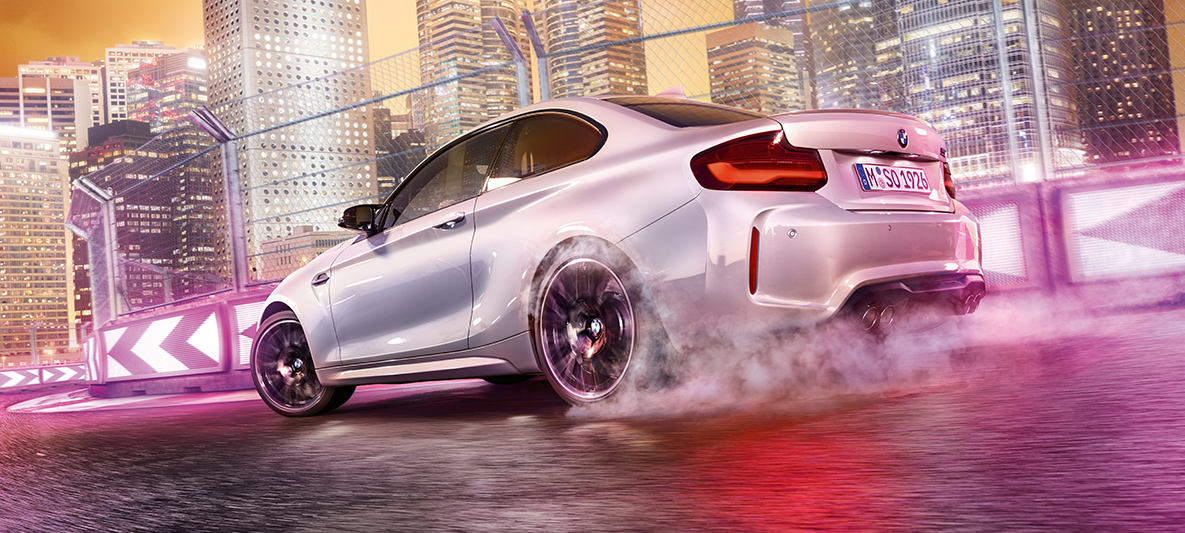 BMW M2 Competition gibt Gas Weißer BMW M2 Competition driftend auf Rennstrecke vor nächtlicher Skyline.