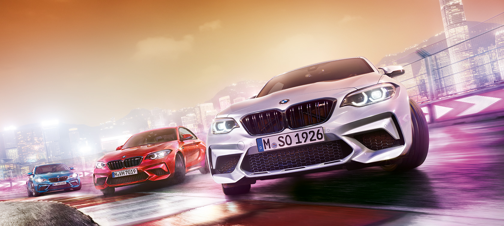 BMW M2 Competition in Aktion Drei BMW M2 Competition driften auf Rennstrecke vor nächtlicher Großstadtkulisse.