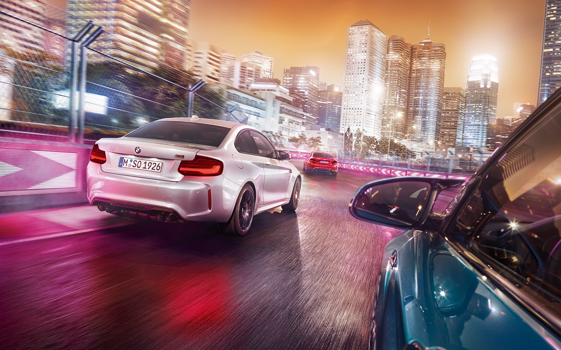 BMW M2 Competition Rückansicht Drei BMW M2 Competition in der Rückansicht fahren auf Rennstrecke vor beleuchteter Skyline.