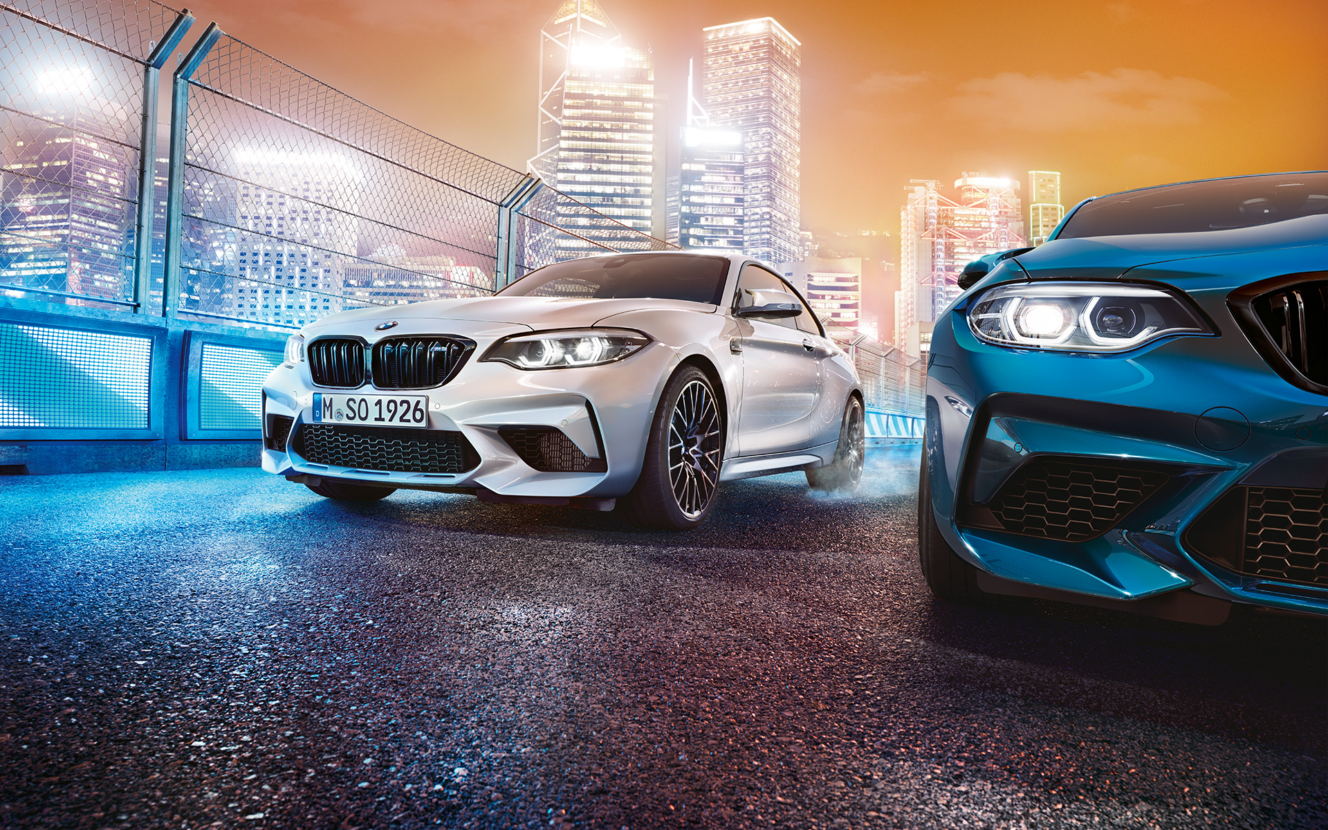 BMW M2 Competition Frontansicht Weißer und blauer BMW M2 Competition in Frontansicht stehend vor nächtlicher Großstadtkulisse.