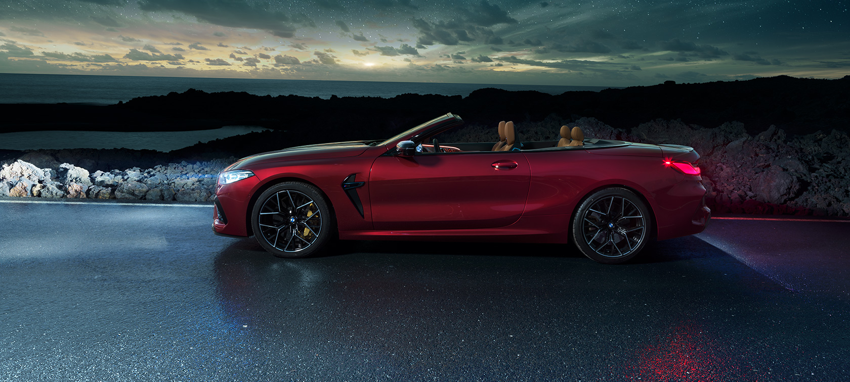 BMW M8 Competition Cabrio, Seitenansicht. BMW M8 Competition Cabrio in Motegi Rot metallic mit geöffnetem Verdeck, Seitenansicht, stehend auf einer Küstenstraße unter Sternenhimmel.
