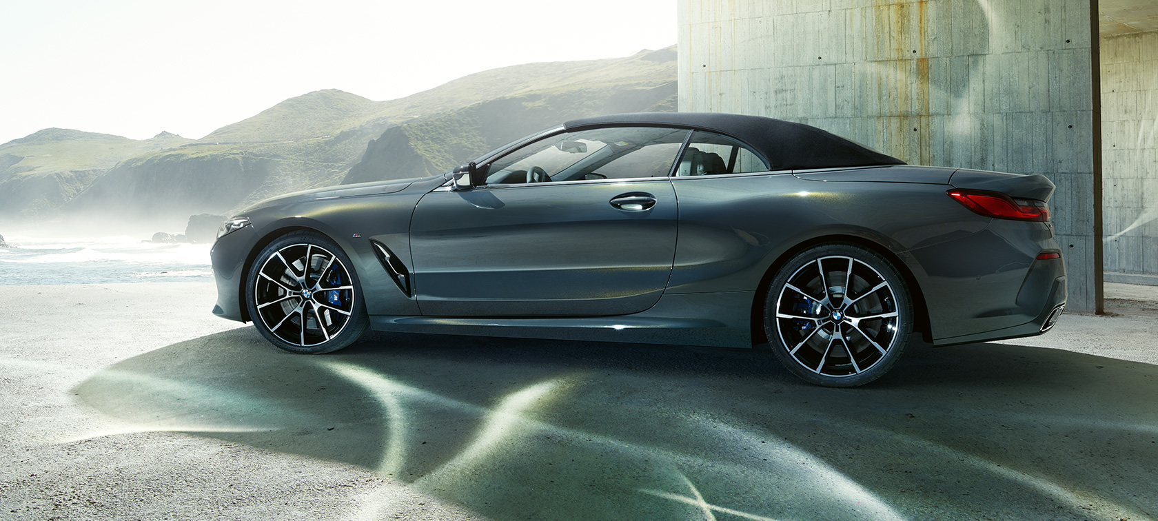 BMW M850i xDrive Cabrio, Seitenansicht. BMW M850i xDrive Cabrio in Dravit Grau metallic mit geschlossenem Verdeck, Seitenansicht vor Hügelkette.