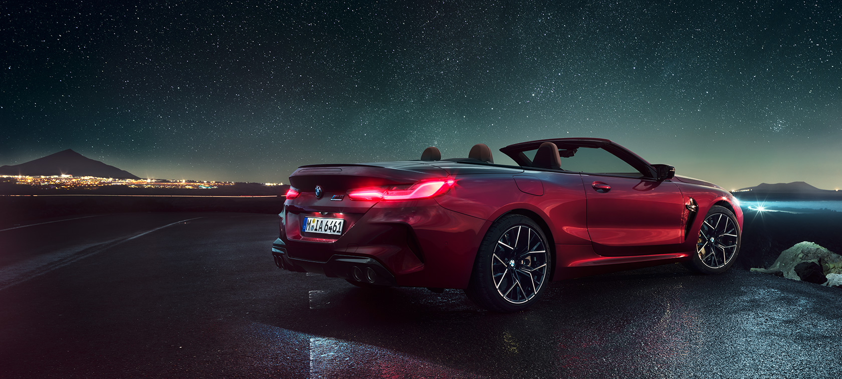 BMW M8 Competition Cabrio, Dreiviertel-Heckansicht. BMW M8 Competition Cabrio in Motegi Rot metallic mit geöffnetem Verdeck, Dreiviertel-Heckansicht, stehend unter Sternenhimmel.