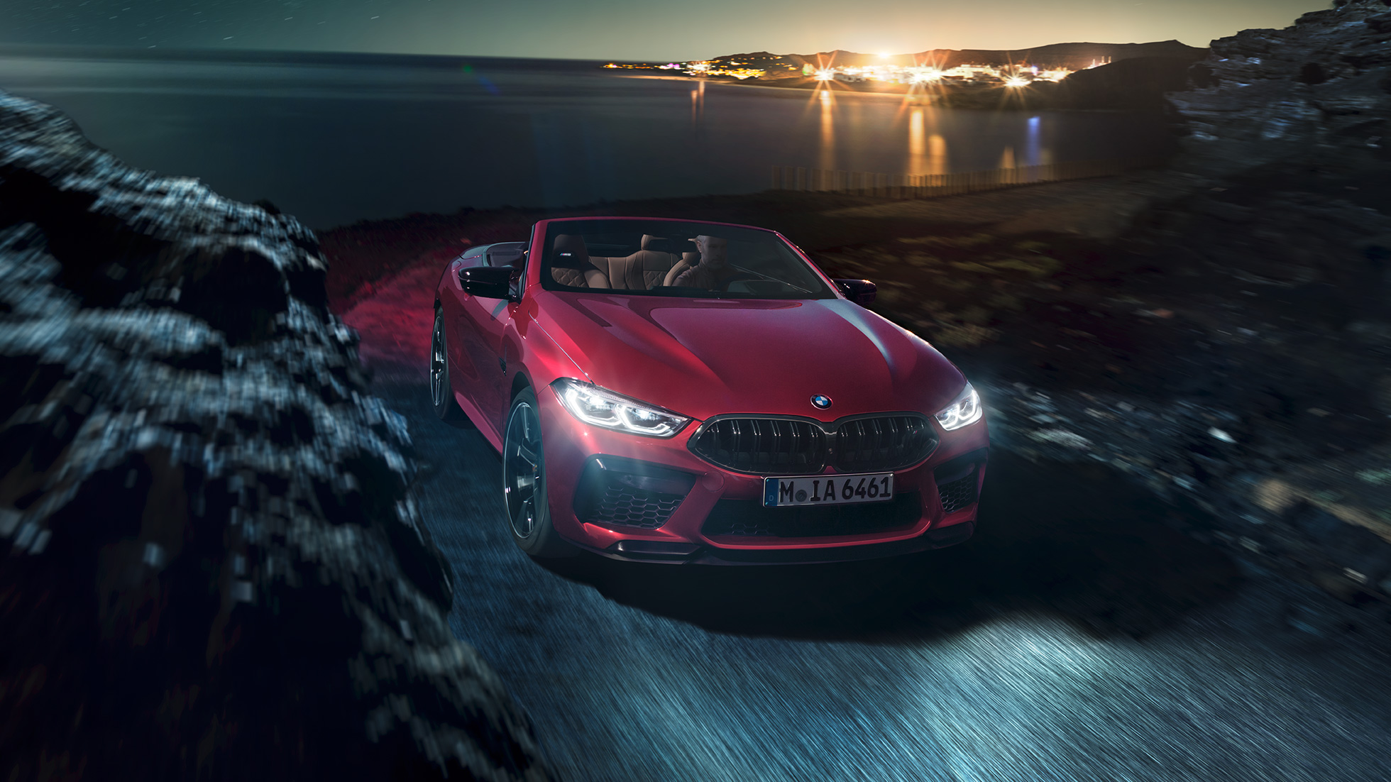 BMW M8 Competition Cabrio, Dreiviertel-Frontansicht. BMW M8 Competition Cabrio in Motegi Rot metallic mit geöffnetem Verdeck, Dreiviertel-Frontansicht, fahrend bei Nacht auf Küstenstraße.