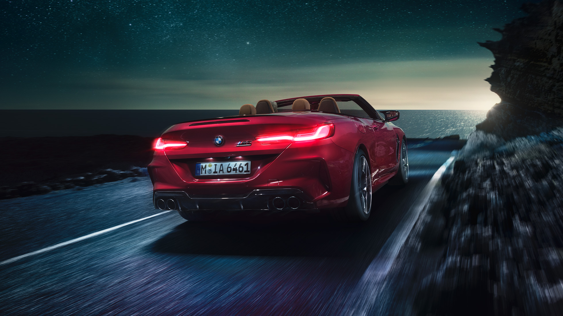 BMW M8 Competition Cabrio, Dreiviertel-Heckansicht. BMW M8 Competition Cabrio in Motegi Rot metallic mit geöffnetem Verdeck, Dreiviertel-Heckansicht, fahrend bei Nacht auf Küstenstraße.