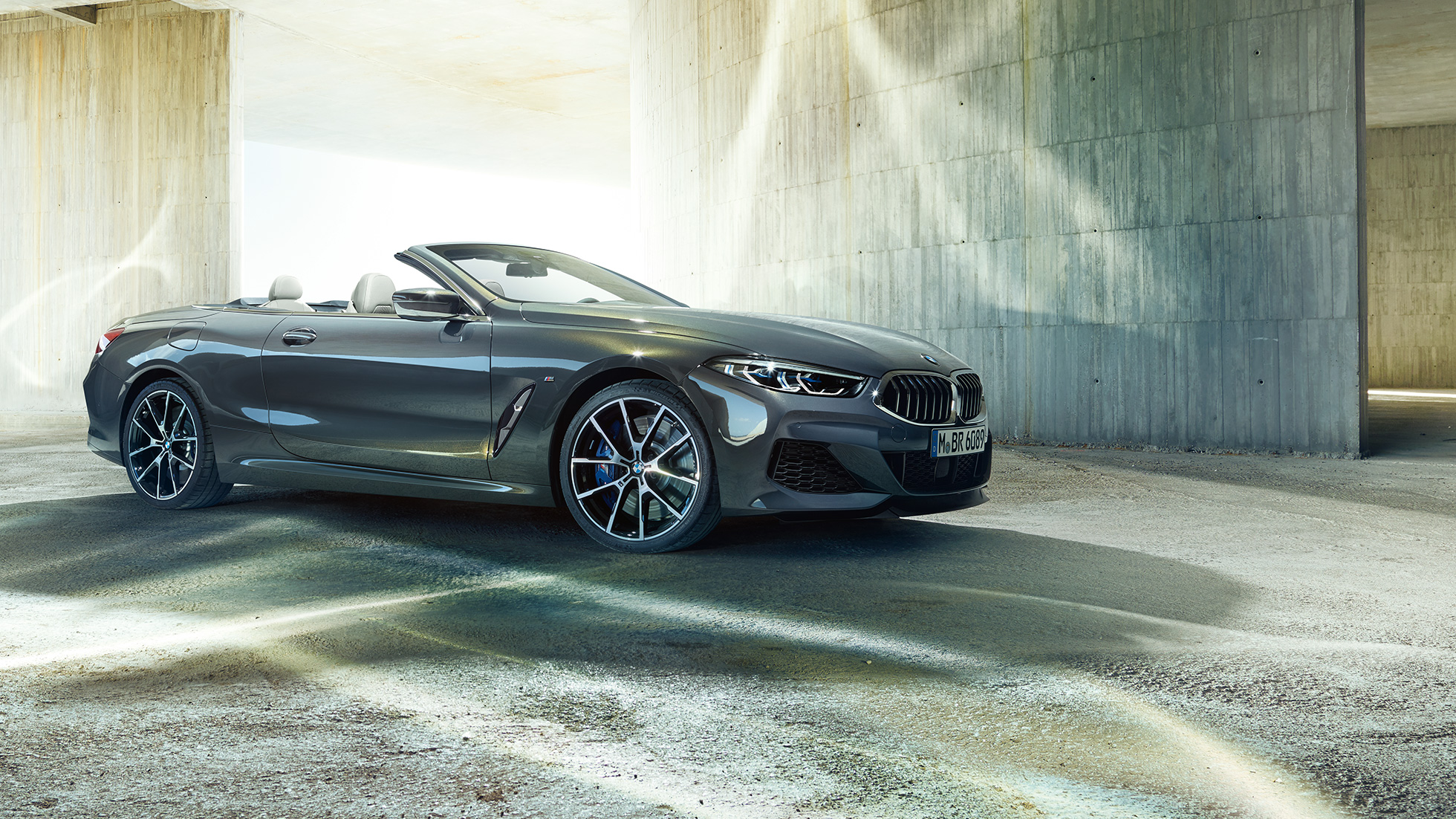 BMW M850i xDrive Cabrio, Dreiviertel-Seitenansicht. BMW M850i xDrive Cabrio in Dravitgrau metallic mit geöffnetem Verdeck, Dreiviertel-Seitenansicht, stehend vor Betongebäude.