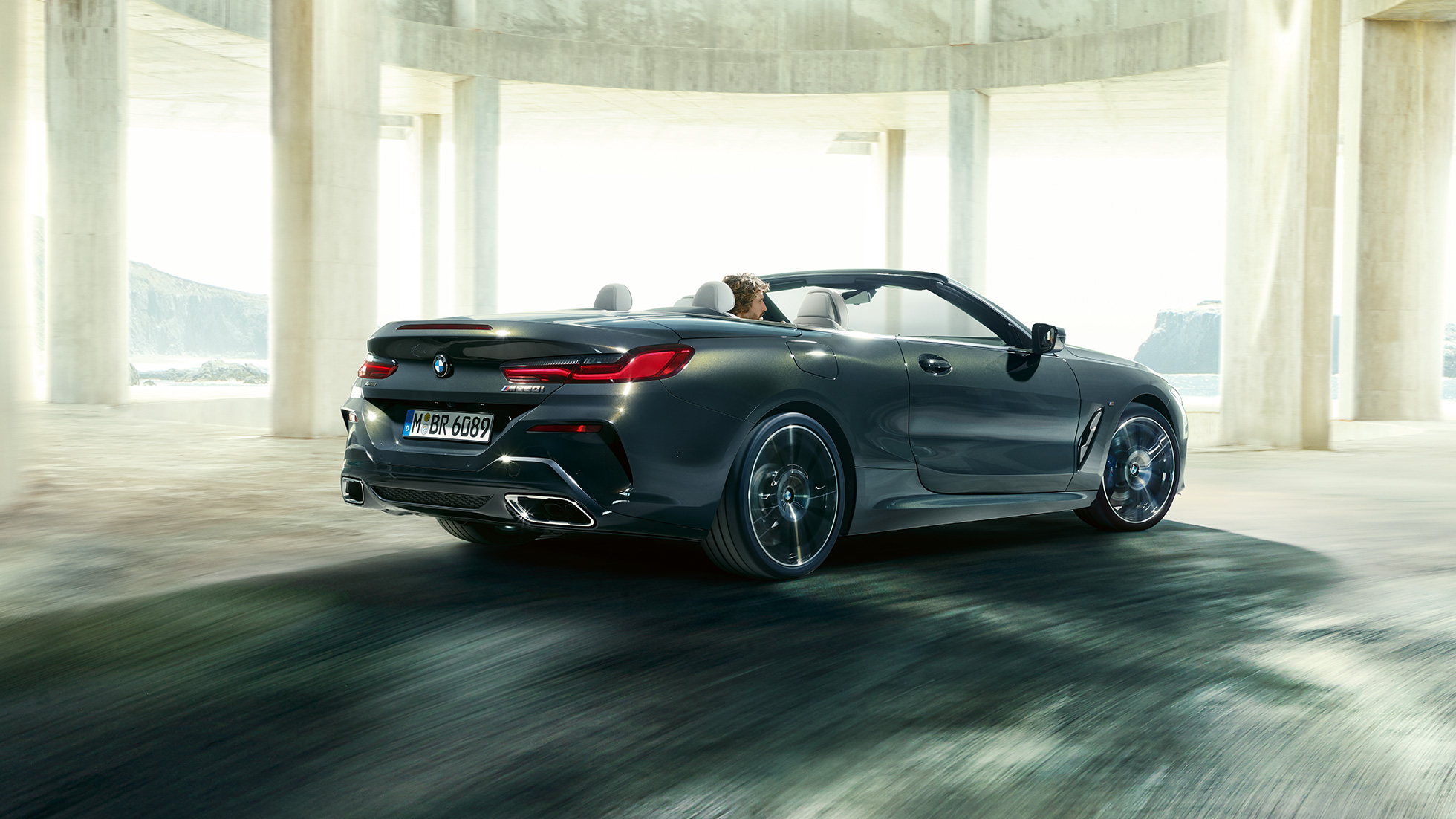 BMW M850i xDrive Cabrio, Dreiviertel-Heckansicht. BMW M850i xDrive Cabrio in Dravitgrau metallic mit geöffnetem Verdeck, Dreiviertel-Heckansicht, fahrend durch Betongebäude.