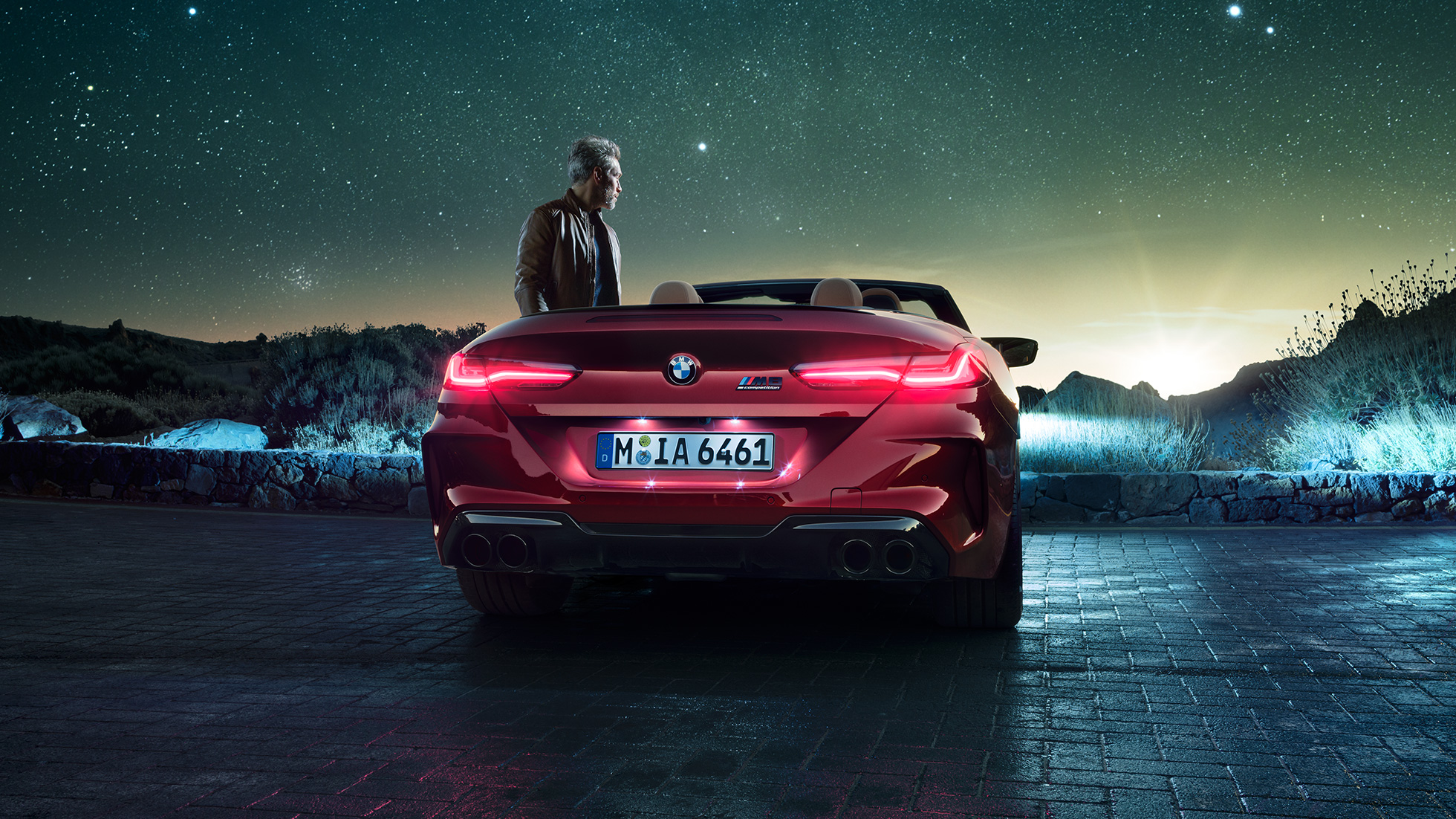 BMW M8 Competition Cabrio, Heckansicht. BMW M8 Competition Cabrio in Motegi Rot metallic mit geöffnetem Verdeck, Heckansicht, stehend mit Mann unter Sternenhimmel.