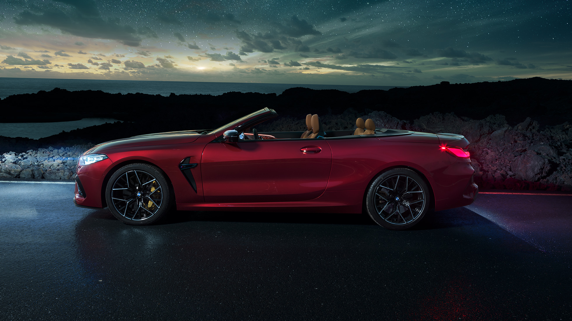 BMW M8 Competition Cabrio, Seitenanicht. BMW M8 Competition Cabrio in Motegi Rot metallic mit geöffnetem Verdeck, Seitenansicht, stehend bei Nacht unter Sternenhimmel.