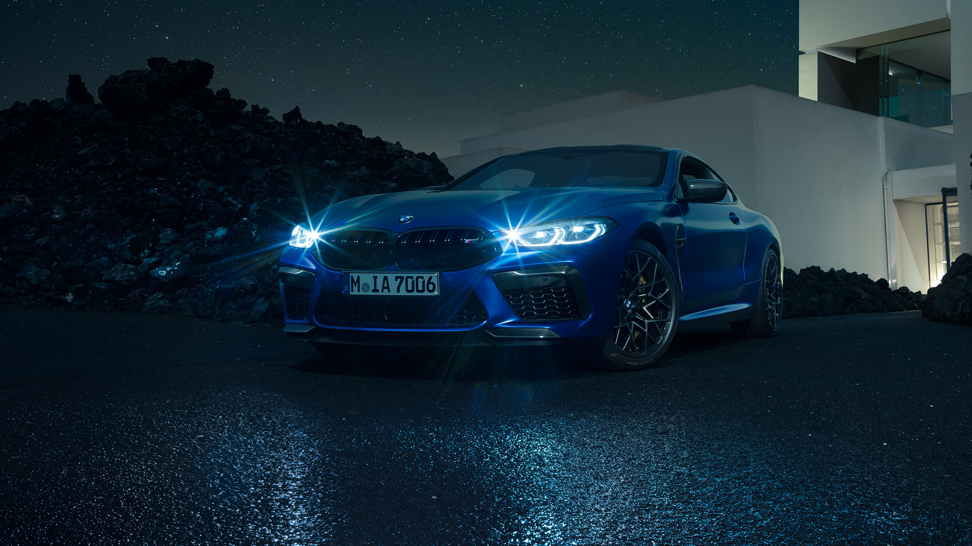 BMW M8 Competition Coupé, Dreiviertel-Frontansicht. BMW M8 Competition Coupé in Marina Bay Blau metallic mit strahlenden LED-Scheinwerfern, stehende Dreiviertel-Frontansicht vor Villa bei Nacht.