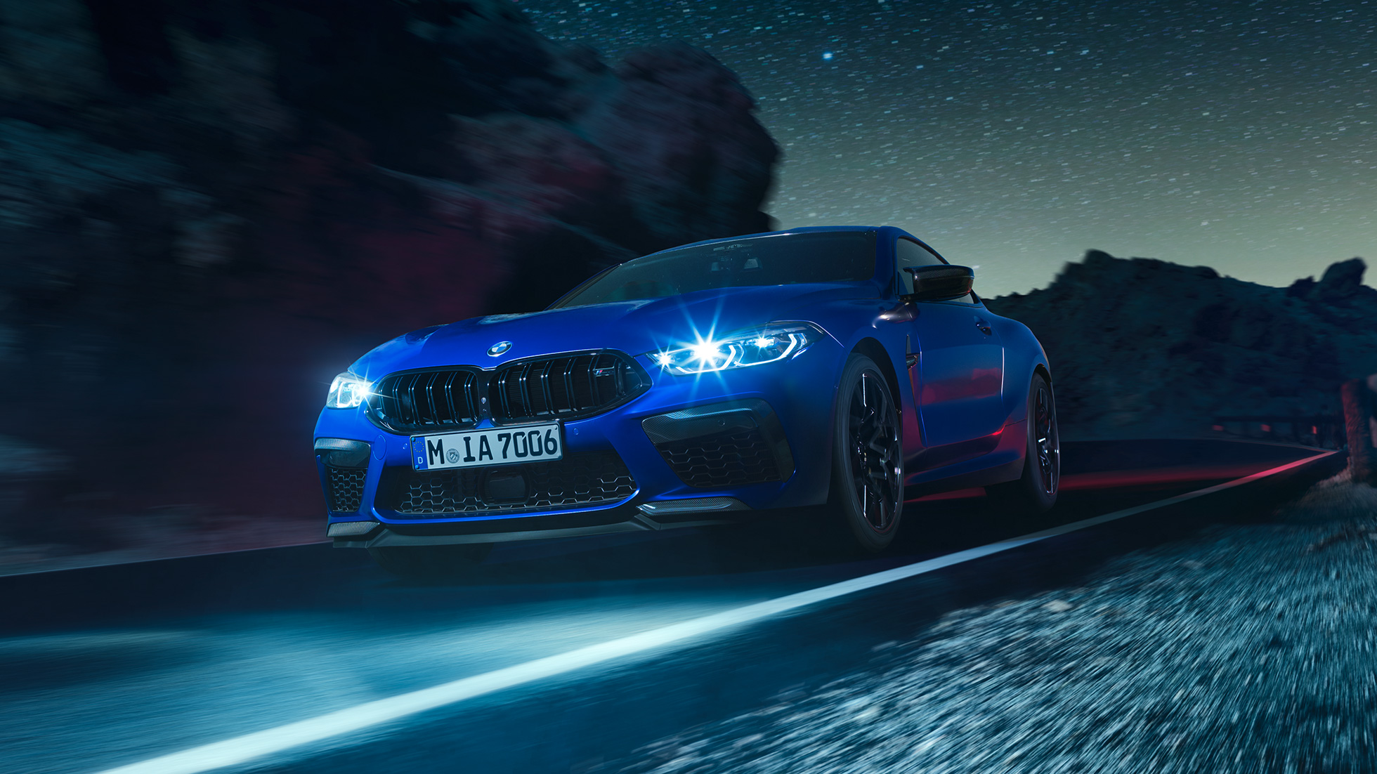 Fahrendes BMW M8 Competition Coupé, Dreiviertel-Frontansicht. BMW M8 Competition Coupé in Marina Bay Blau metallic mit strahlenden LED-Scheinwerfern, Dreiviertel-Frontansicht, fahrend bei Nacht.