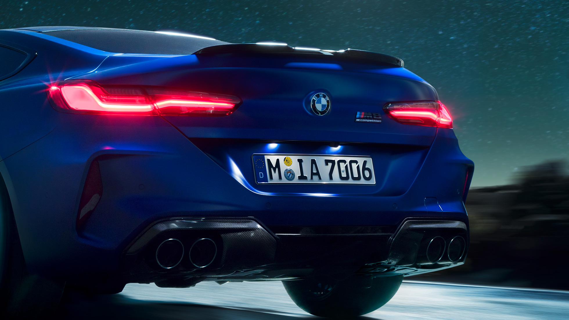 M Diffusor in Carbon Schwarz. BMW M8 Competition Coupé in Marina Bay Blau metallic, Nahaufnahme Heck bei Nacht mit M Diffusor in Carbon Schwarz.