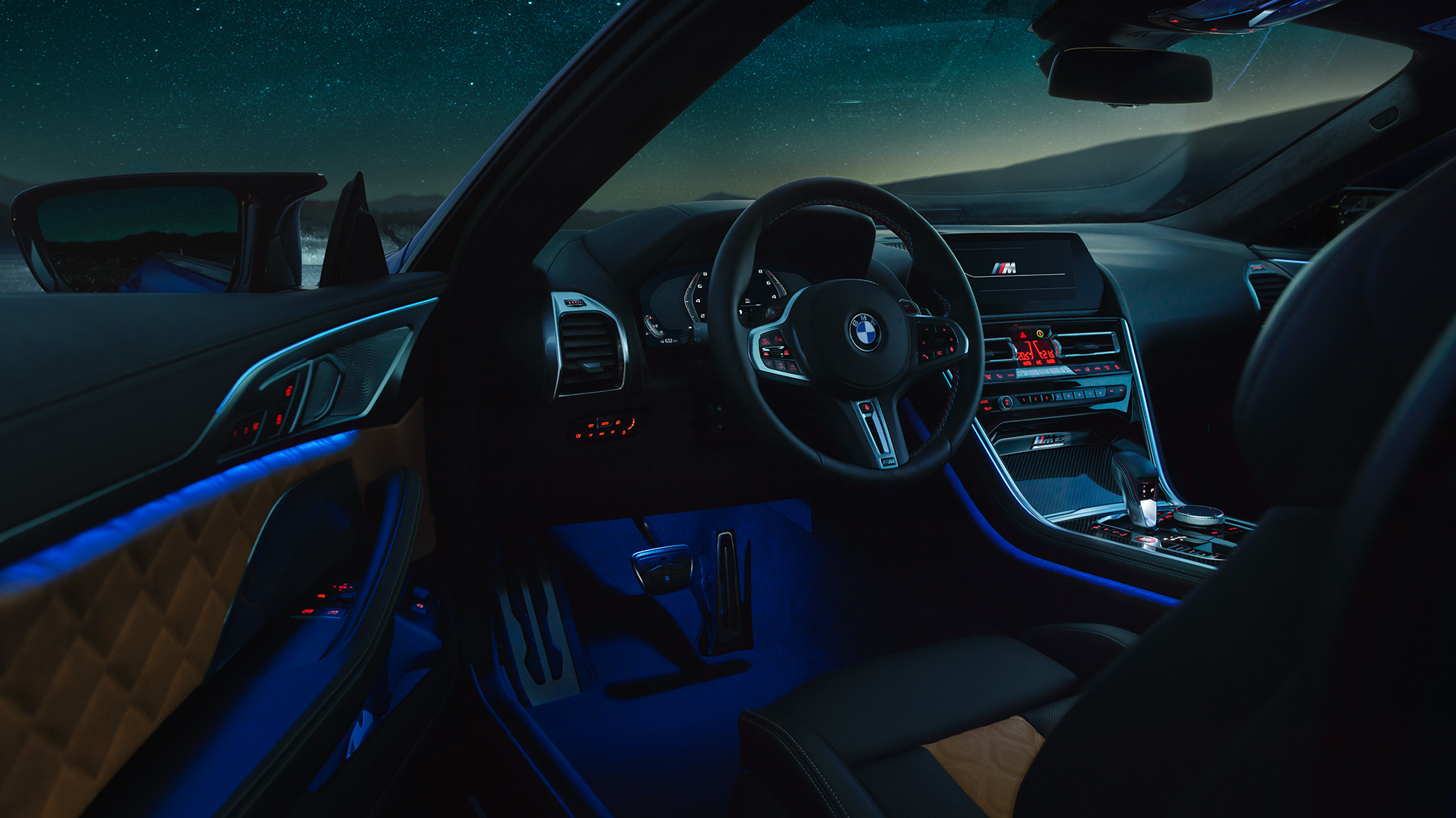 Interieur BMW M8 Competition Coupé. BMW M8 Competition Coupé, Interieur bei Nacht mit M Lederlenkrad und blauem ambienten Licht bei geöffneter Fahrertür.