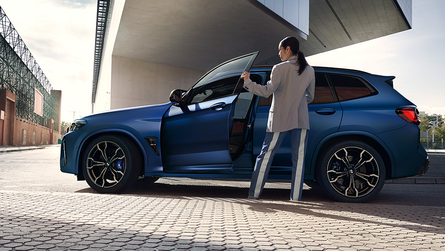 BMW X3 M Competition F97 LCI 2021 Seitenansicht mit geöffneter Tür und weiblichem Model BMW X3 M Competition F97 LCI Facelift 2021 Frozen Marina Bay Blau metallic Seitenansicht mit geöffneter Tür und weiblichem Model