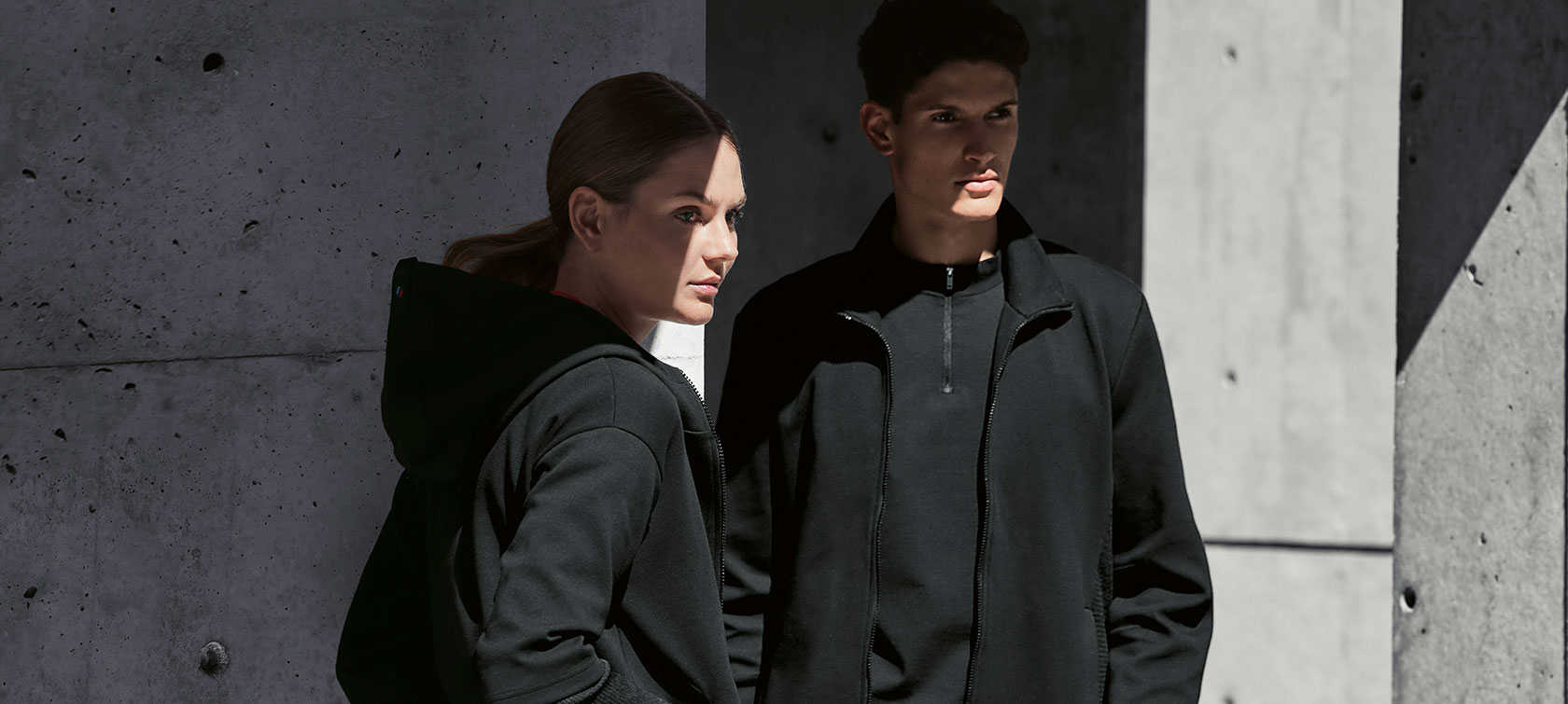 BMW M Sweatjacke für Damen und BMW M Sweatjacke für Herren. Ein Paar steht im Schatten. Eine Frau präsentiert die BMW Sweatjacke für Damen und ein Mann trägt die BMW Sweatjacke für Herren.
