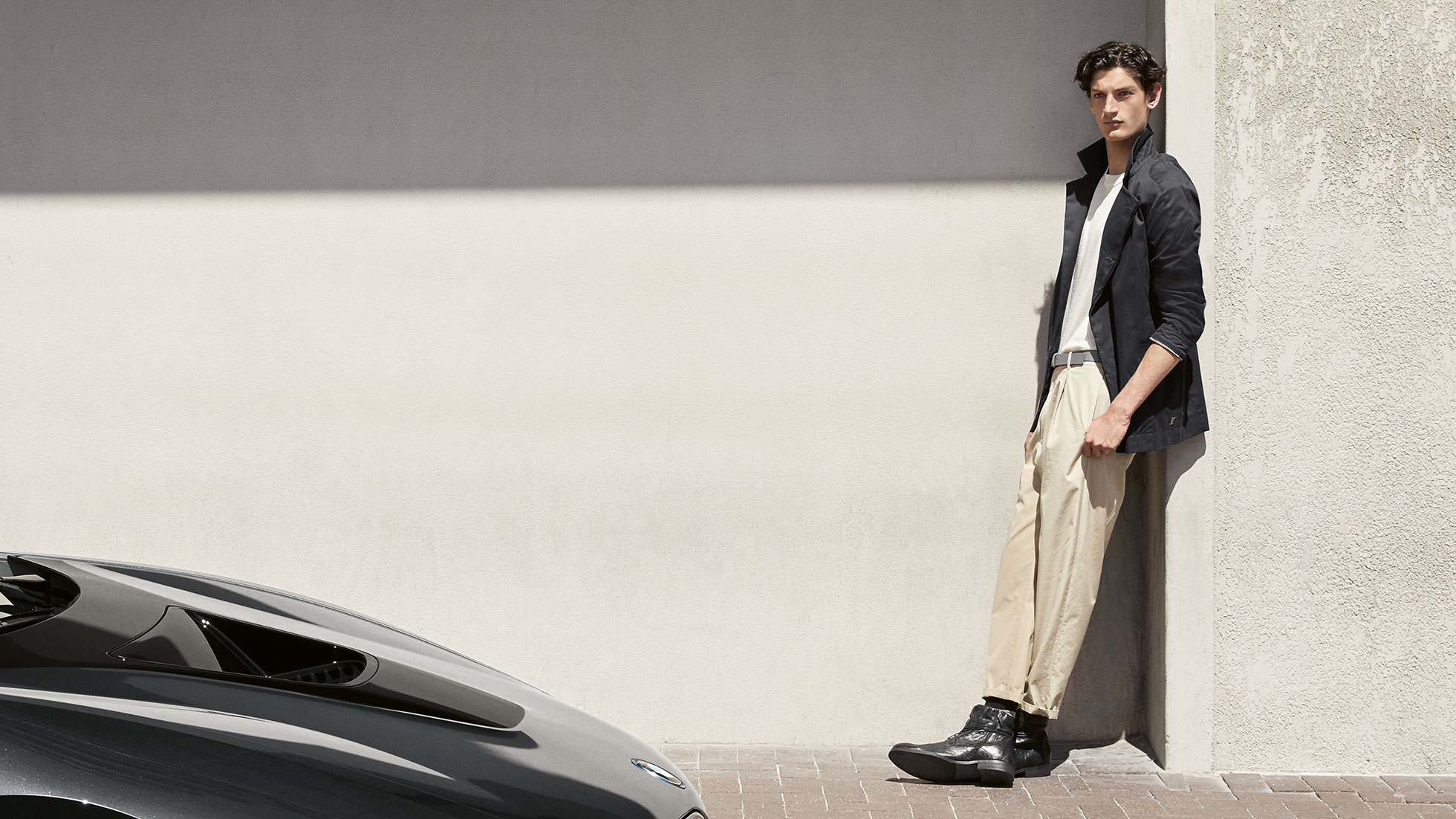 BMW i Collection. Das Bild zeigt einen Mann, der sich in der BMW i Jacke aus der BMW i Collection an eine Wand lehnt.