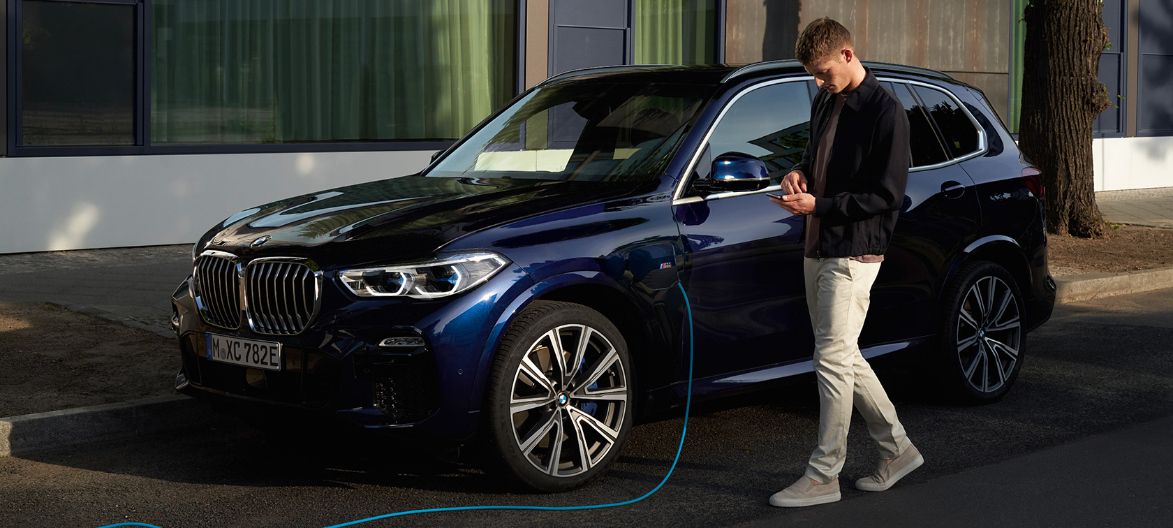 Mann nutzt My BMW App auf dem Smartphone Mann nutzt My BMW App von BMW ConnectedDrive auf dem Smartphone während sein BMW X5 Plug-In Hybrid aufgeladen wird