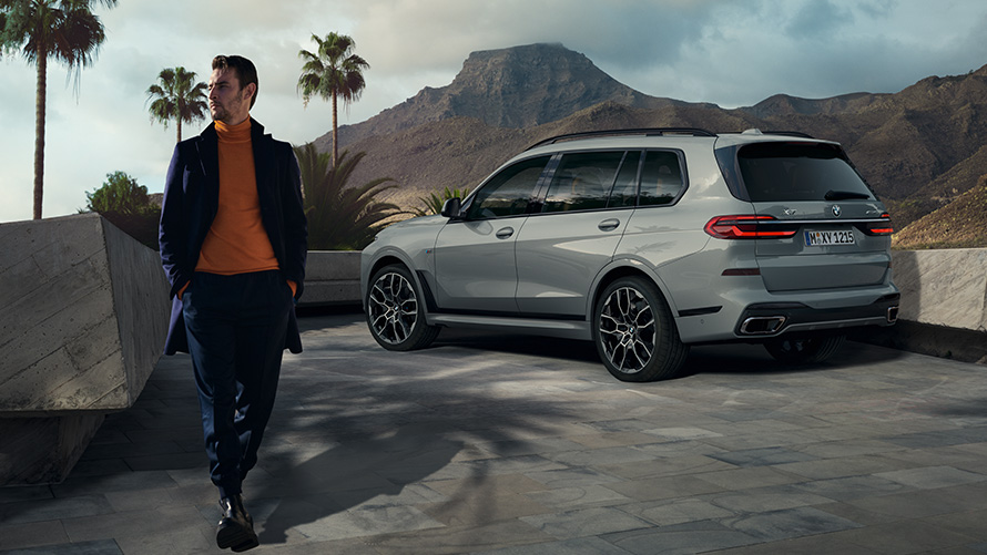Mann vor geparktem BMW X7 (G07) Remote Control Parking Remote Control Parking Mann vor geparktem BMW X7 (G07) Dreiviertel-Heckansicht unter Palmen mit Berglandschaft im Hintergrund