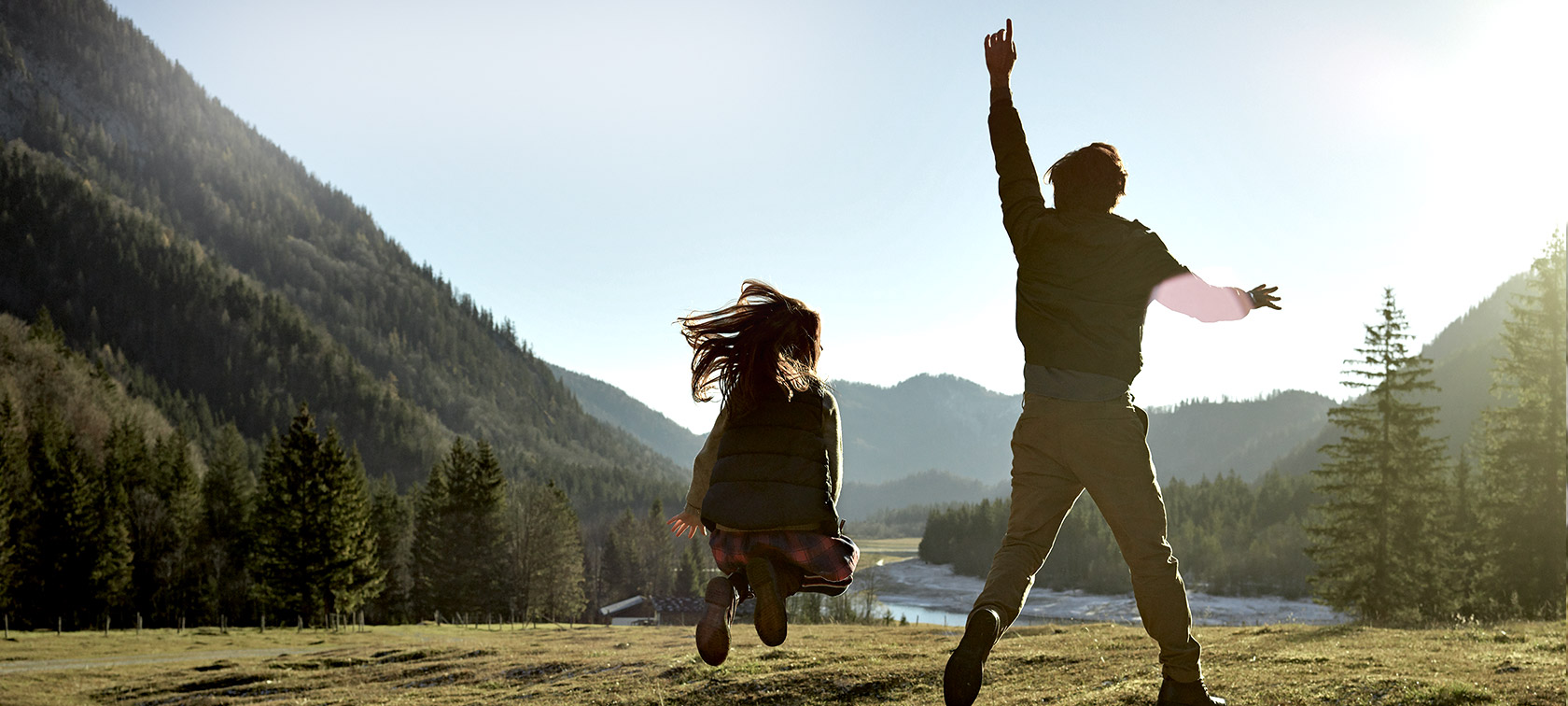Mann und Mädchen auf Wiese in Bergtal, Rückansicht Mann und Mädchen springend auf Wiese in Bergtal, Rückansicht