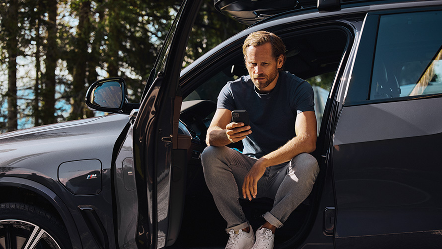 Mann sitzt in einem BMW und guckt auf sein Smartphone BMW Frühlings- und Sommerzubehör Mann sitzt in einem BMW und guckt auf sein Smartphone