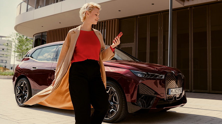My BMW App My BMW App BMW iX i20 Frau gehend vor einem BMW Fahrzeug mit Smartphone in der Hand