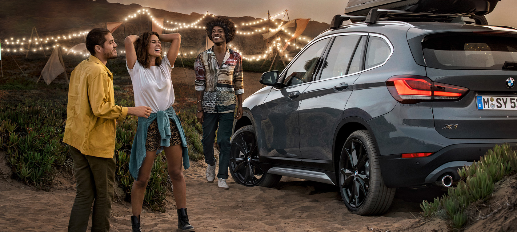 Jubelndes Trio in BMW X1 mit BMW Angebote Sommer Glückliches Trio in BMW X1 Mineralgrau ausgestattet mit BMW Angebote Sommer Dreiviertel Heckansicht auf Strand