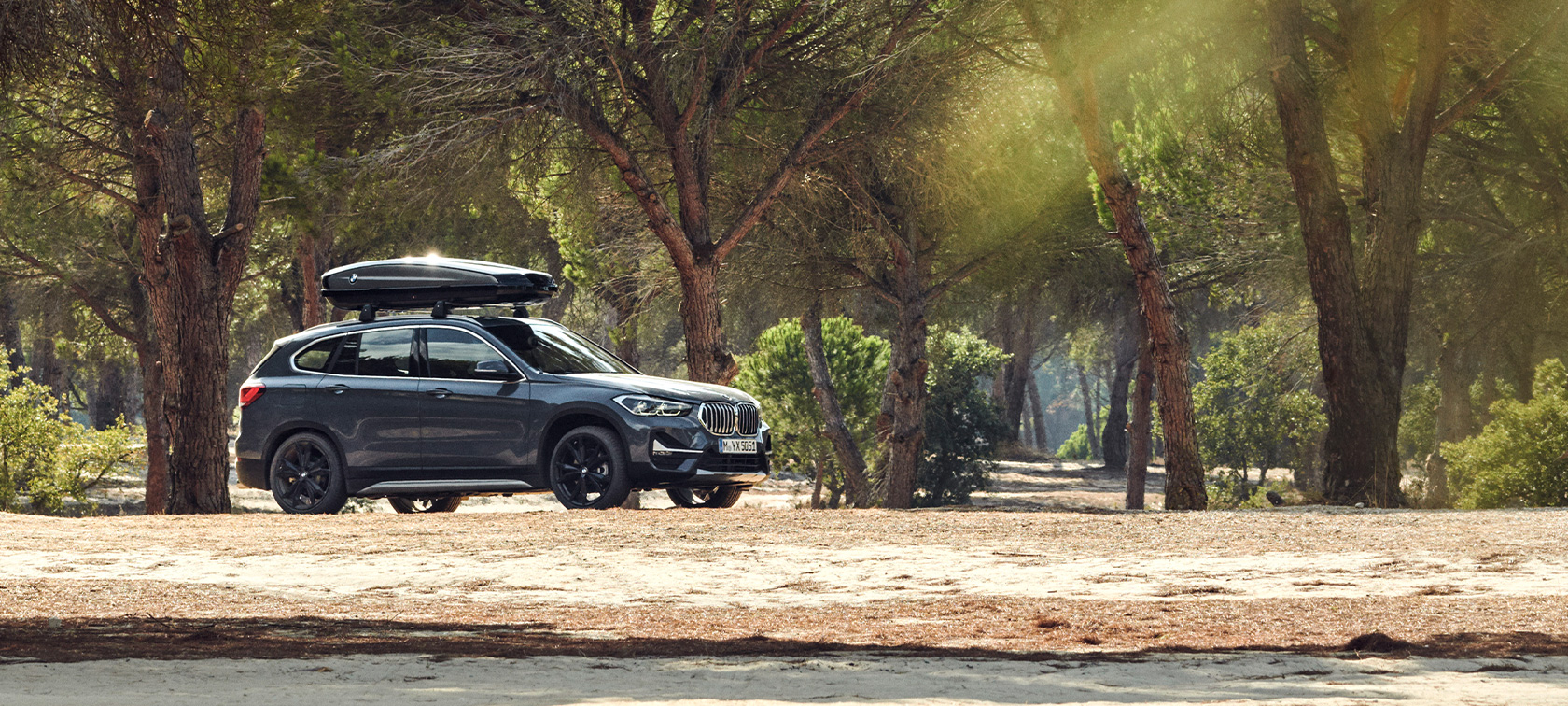 Seitenansicht BMW X1 mit Dachbox BMW Angebote Sommer Seitenansicht BMW X1 Mineralgrau mit BMW Dachbox stehend offroad in Wald BMW Angebote Sommer