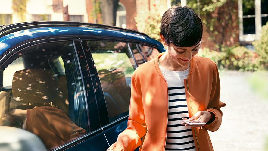 BMW Angebote Sommer BMW Teleservices BMW Angebote Sommer BMW Teleservices Frau steht vor BMW und nutzt Smartphone