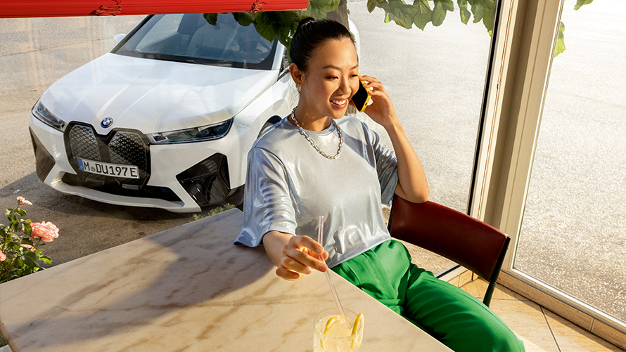 Voraussetzungen Proactive Care Proactive Care, Check-up & Wartung Voraussetzungen Proactive Care Proactive Care, Check-up & Wartung Frau sitzend am Tisch mit Smartphone in der Hand und mit BMW Fahrzeug im Hintergrund