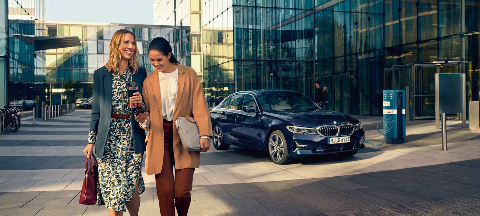 Zwei gut gelaunte Frauen durchqueren einen Bürokomplex. Seitlich steht ein Elektro BMW beim Aufladen. Zwei gut gelaunte Frauen durchqueren einen Bürokomplex. Seitlich steht ein Elektro BMW beim Aufladen.