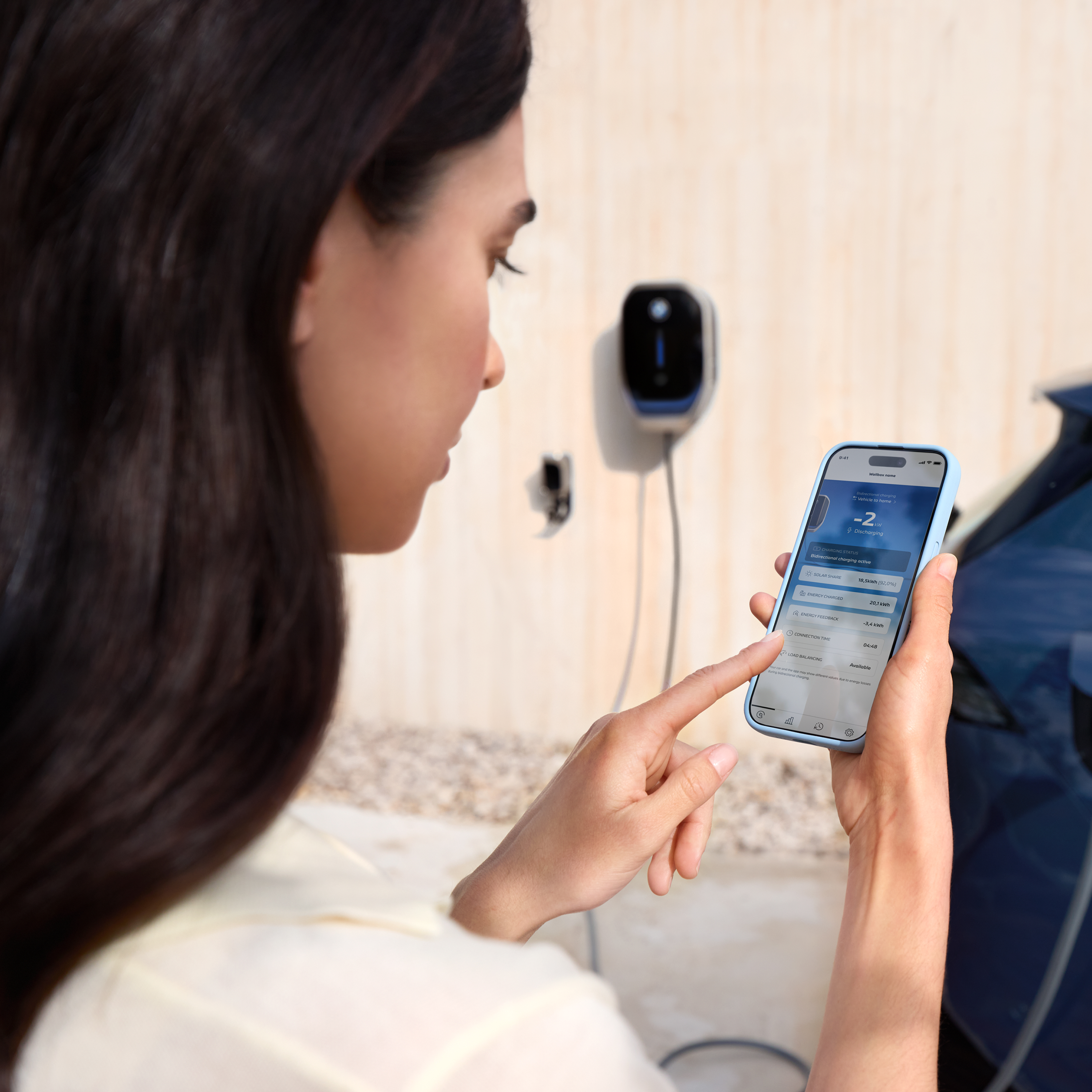 Eine Frau blickt auf Ihr Smartphone, im Hintergrund ist eine BMW Wallbox