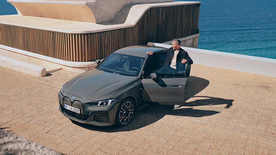 Eine Frau lehnt sich lässig gegen einen vollelektrischen BMW. Ein Mann steigt in einen BMW, der auf einem Parkplatz eines Gebäudes an der Küste abgestellt ist.