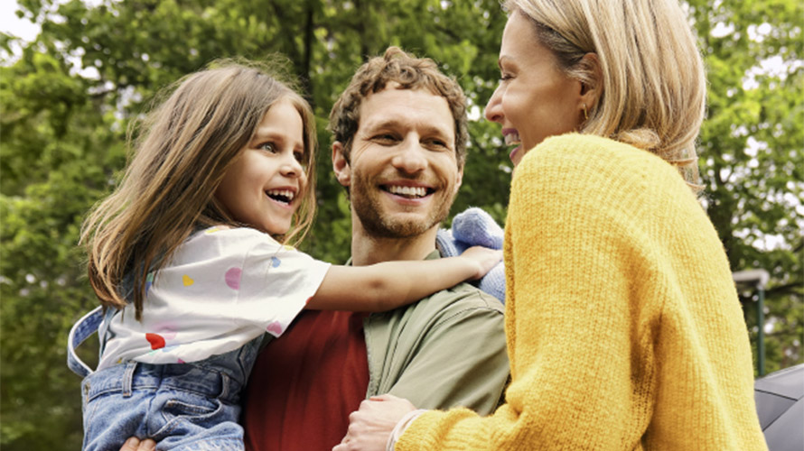 Eine lachende Familie. Die junge Tochter wird vom Vater im Arm gehalten.